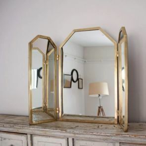 A gold dressing mirror with 3 panels placed on a natural wood sideboard.