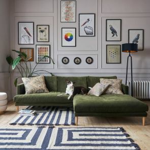 A wide shot of a living room with a green velvet mid-century corner sofa with a dog sat upon it, two bold striped rugs in white and navy lay in front of the sofa, a gallery wall frames behind.