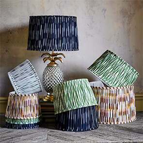 A stack of ikat lamp shades in a variety of colours from green, navy and yellow surround a silver and gold pineapple table lamp topped with a navy blue ikat lamp shade.