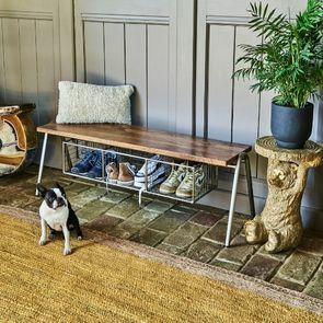 A stone-bricked hallway with a French bulldog sitting upon a jute woven rug. A shoe rack bench filled with dirty trainers sits behind. 