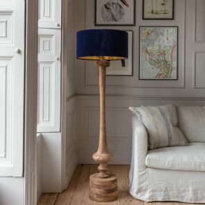 A hand turned natural wood floor lamp topped with a navy-blue lamp shade surround by a natural linen sofa and white shutter windows.