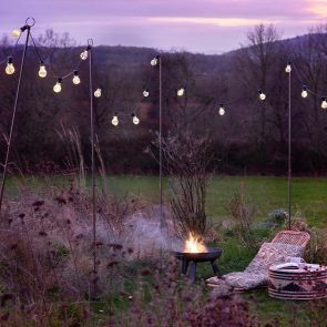 Dusk in a garden meadow with a sun lounger next to a lit fire pit, framed by festoon string lights.