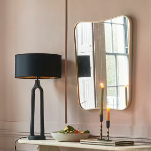 A rounded rectangular gold rimmed wall mirror is hung behind a console table which as a sculptural black table lamp and green candles in rustic black candle holders.