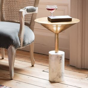 A sculptural gold and marble side table has a singular book placed upon it with a wooden and grey velvet armchair to the left-hand side.