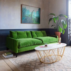 A velvet green sofa contrasted against a diamond patterned white and blue rug with an abstract green and teal painting behind.