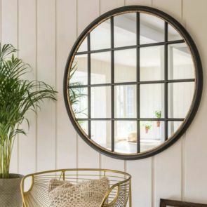 A large circular window pane with a rustic black finish inlayed with gold is hung on a white panelled wall, a gold accent chair with a linen cushion placed on it.