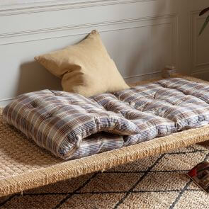 A lavender and brown striped bed roll is folded on a woven day bed with a beige linen square cushion placed on top of it.