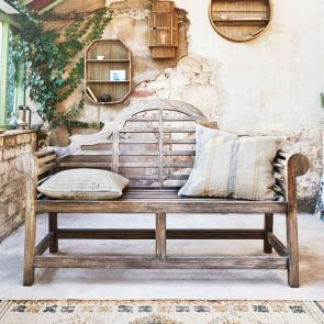 A teak wooden bench decorated with natural linen square cushions surround by bamboo shelves holding treasured items.