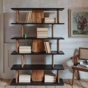 A contrasting black and mango wood open shelving unit adorned with stacks of books and a mid-century rattan backed chair placed next to the shelf.