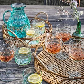 Two rattan trays, laid on a grey teak dining table, filled with clear and blue glasses and tumblers filled with summer drinks.
