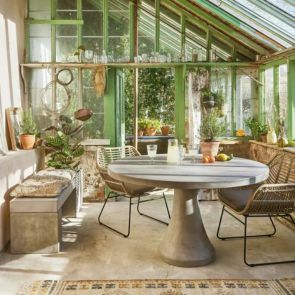 A concrete round dining table and rattan dinging chair in a rustic green conservatory with a concrete bench covered in block printed seat pads to the side