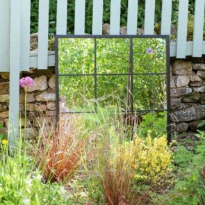 A rectangular window mirror resting against a stony wall reflecting the green foliage of the garden.