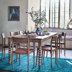 A large mango wood dining room table with large bobbin legs surrounded by 7 teak dining chairs placed upon a large teal woven rug.