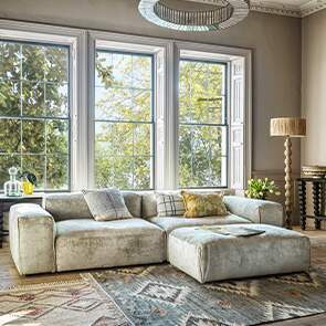 A large modular chenille sofa in front of large sash windows decorated with a bobbin floor lamp and geometric rug.