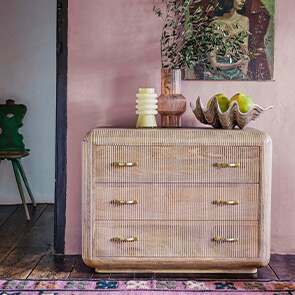 A mid-century mango wood chest of drawers with whitewashed grooves standing against a dusty pink wall decorated with a large clam shell and sculptural candle holders.