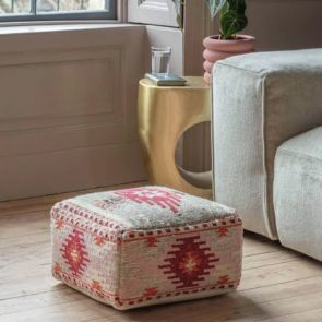 A handwoven pouffe with rich reds on a cream background is surrounded by a linen sofa and a sculptural gold side table.