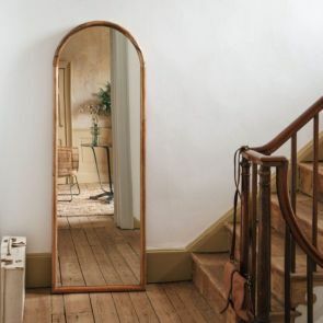 A natural wood framed floor length mirror placed on a landing with the stairs continuing upwards.